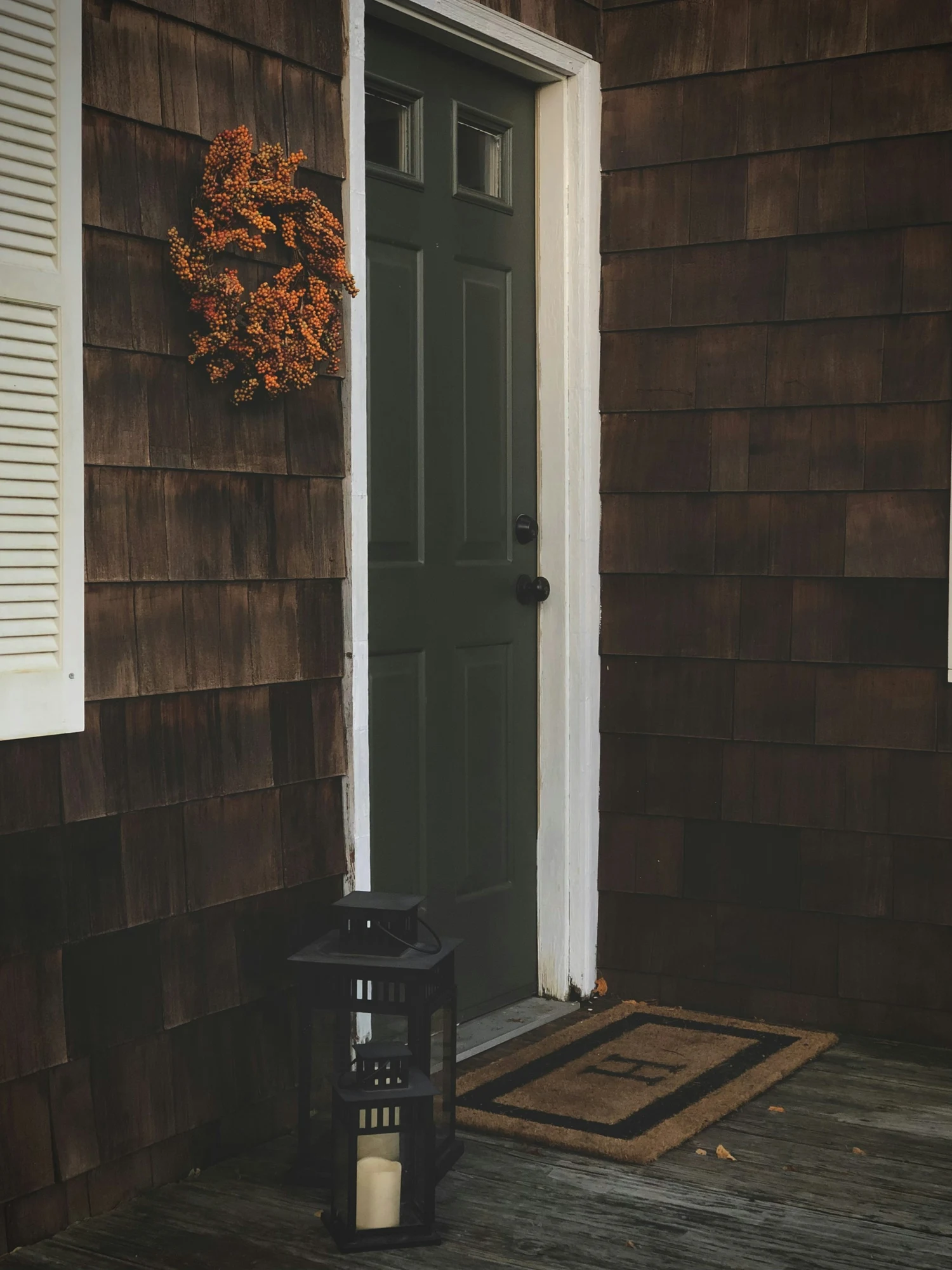 Real green front door beside an orange wreath on cedar siding, with two black lanterns on the porch